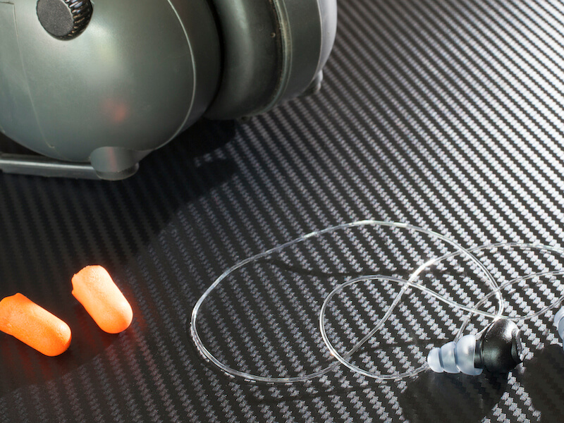Examples of hearing protection - grey earmuffs, orange foam plugs, clear wire earplugs - on a silver table.