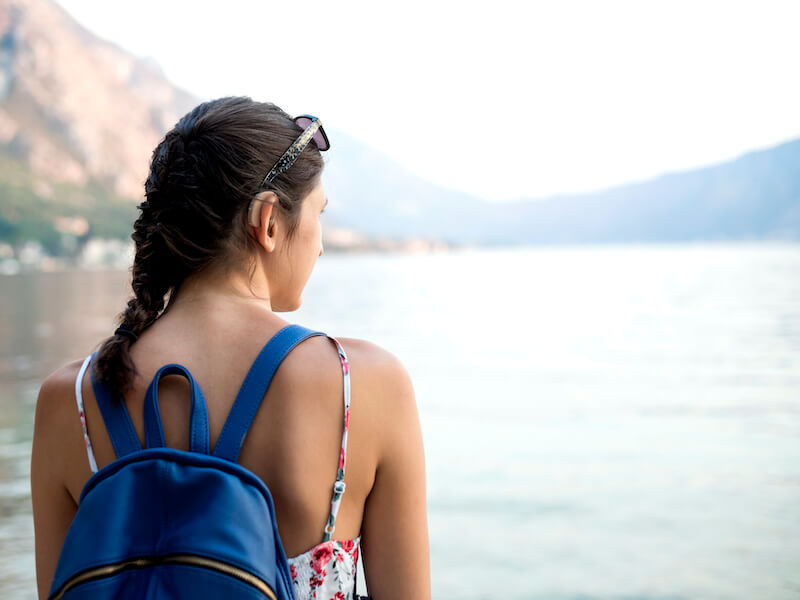 Woman with hearing aids in her ears wearing a backpack overlooking a lake on a summer day.