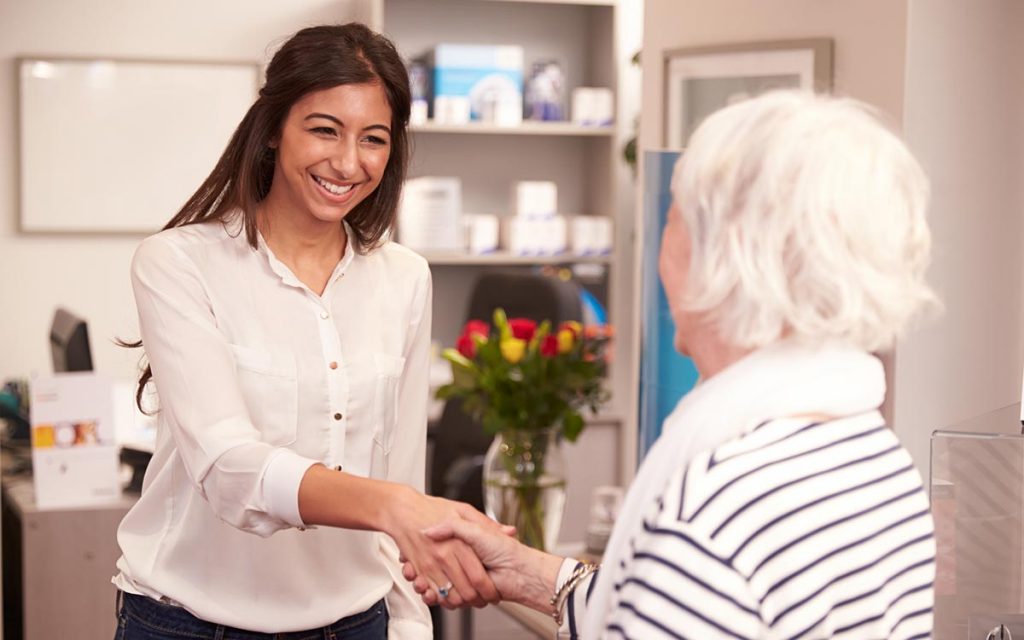 A female Audiologist greeting an elderly woman in the office for a hearing aid fitting.
