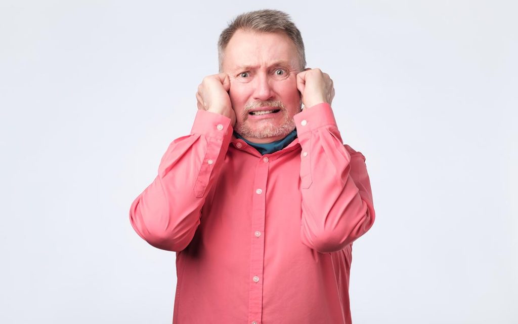 Senior man in red shirt plugging ears with fingers suffering from Tinnitus.