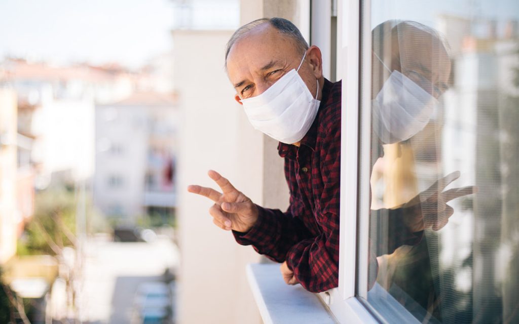 Senior man in medical mask on window breathing fresh air from quarantine not losing his hearing aids from face mask.