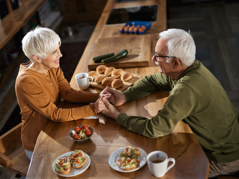 High angle view of happy senior couple holding hands and communicating about hearing loss during morning time in dining room.