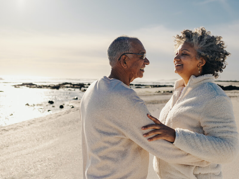 Mature couple wearing hearing aids smiling and dancing together on beach sand. Happy elderly couple having a good time next to the ocean. Cheerful senior couple enjoying their retirement days together.