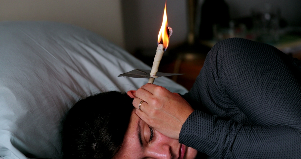 Woman lying in bed holding burning earwax candle cone.