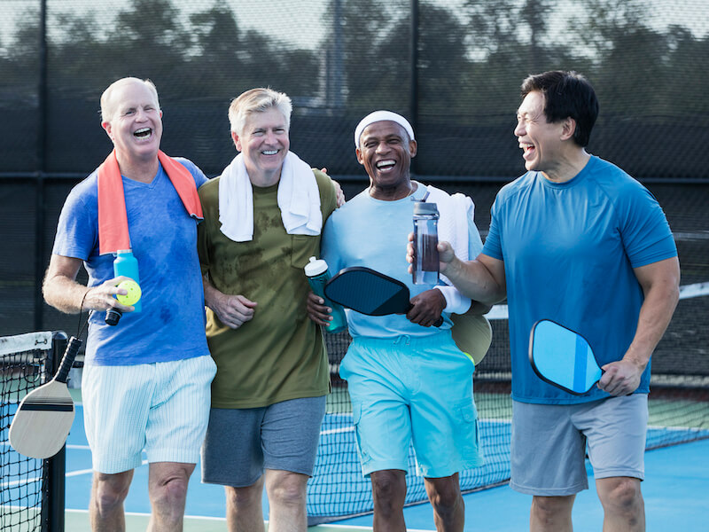4 older, sweaty gentlemen laughing on the tennis court after playing a game of pickle ball