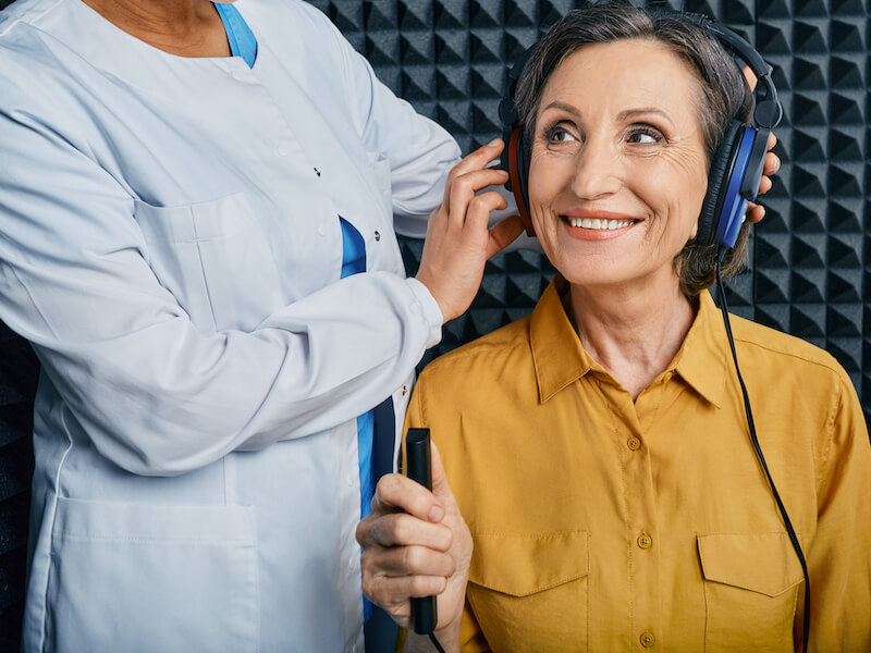 Older woman wearing a yellow button shirt is sitting in a sound booth getting fitted with headphones for her hearing test