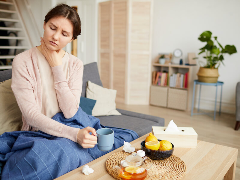 Woman sitting on couch clutching her sore throat while drinking herbal honey and lemon tea