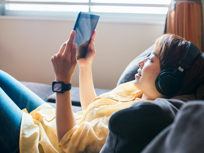 adult woman relaxing on a sofa using a digital tablet and wearing headphones and listening at an unsafe volume.