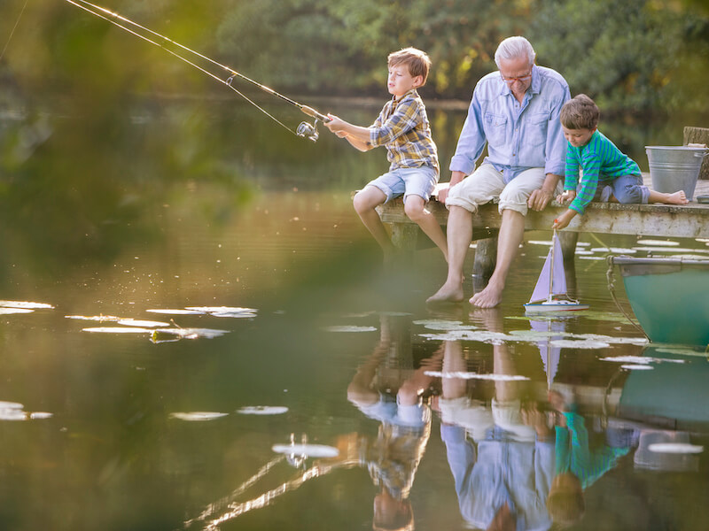 Grandfather wearing hearing aids and enjoying his grandsons fishing and playing with toy sailboat at a lake.