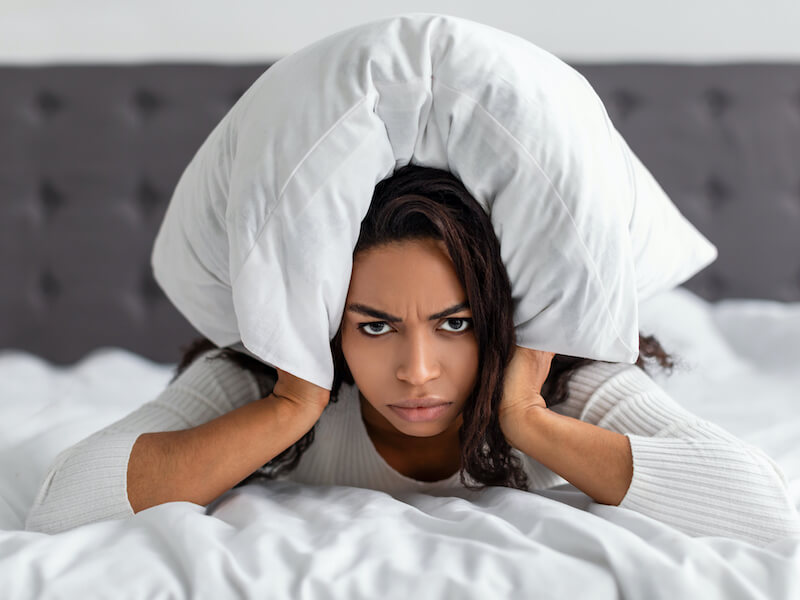 Upset woman suffering from tinnitus laying in bed on her stomach with a pillow folded over the top of her head and ears.