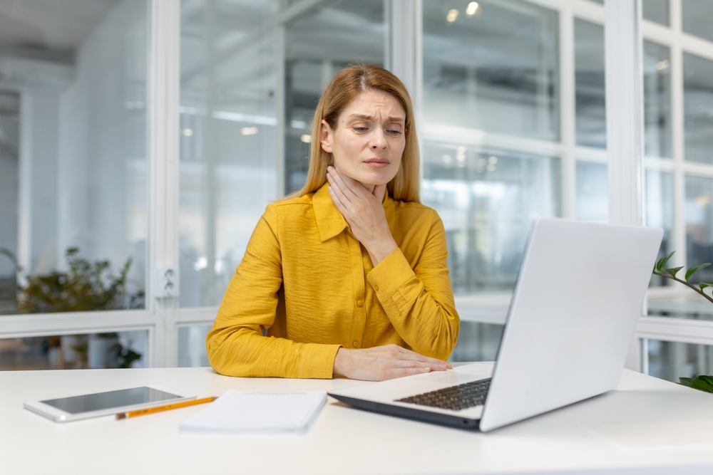 woman in a bright yellow blouse looks uncomfortable and holds her throat while working on a laptop.