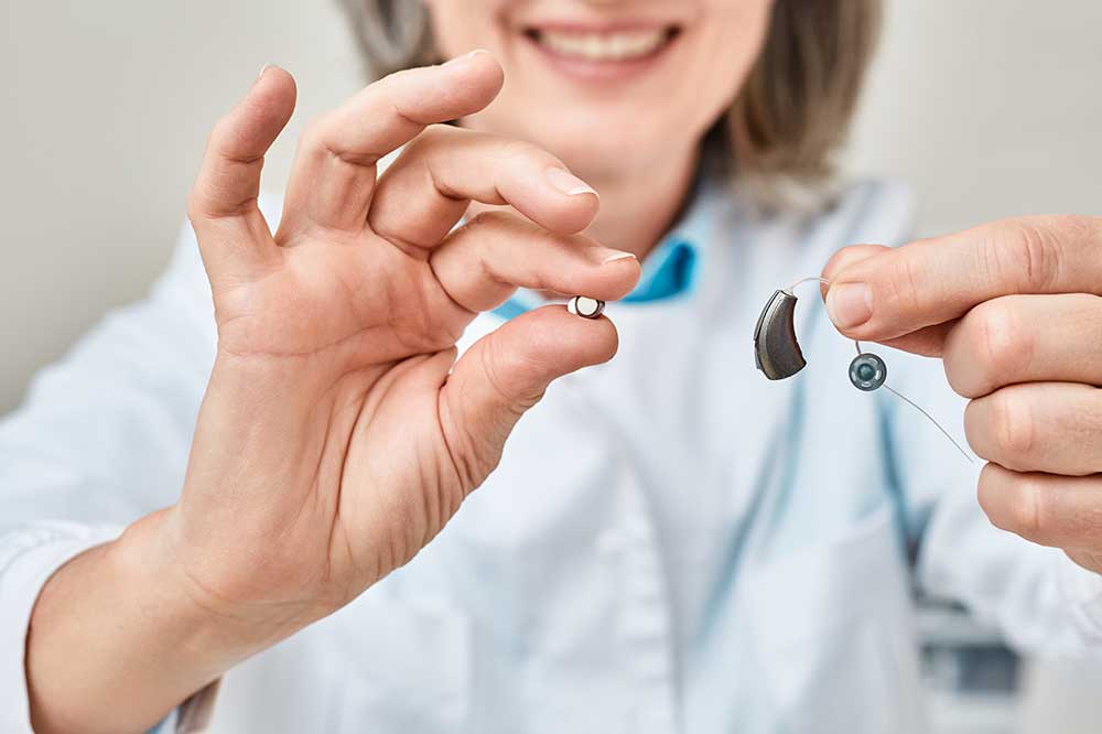 Smiling hearing care professional holding up a hearing aid battery and a receiver-in-canal hearing aid at High Point Audiological in High Point, NC