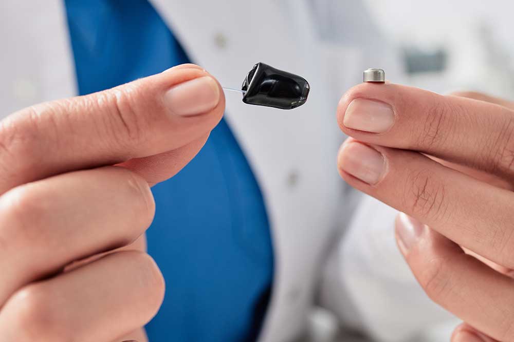 Hands holding a tiny in-canal hearing aid and a button battery, showing hearing aid battery care at Illini Hearing in Champaign, IL