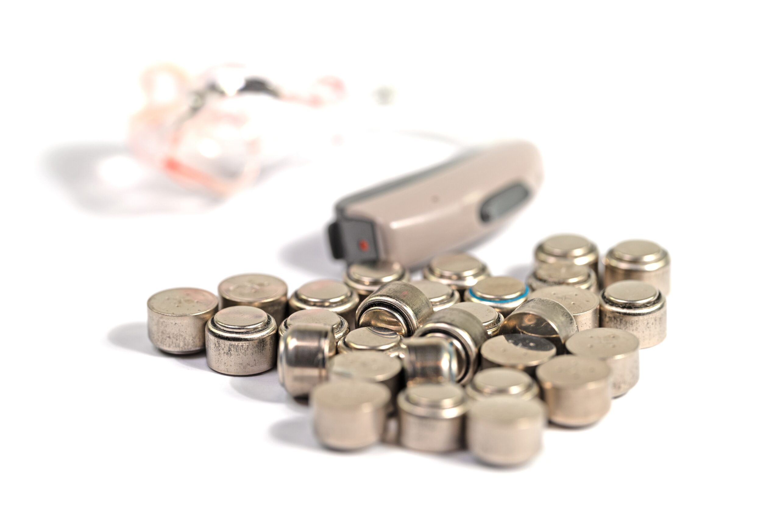 Pile of small disposable hearing aid batteries in front of a behind-the-ear hearing aid, for battery tips from The Hearing Place in Calgary, AB