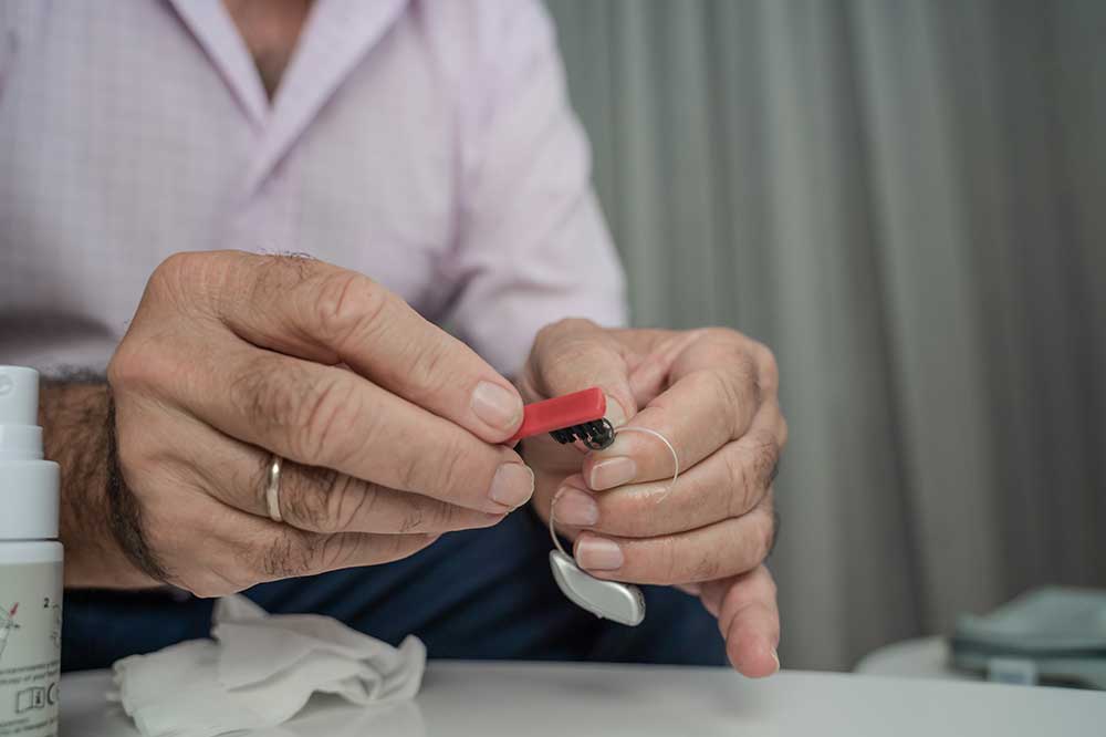 Man brushing a receiver-in-canal hearing aid with a small red brush, a daily cleaning step explained by Aspire Hearing Solutions in Lakeland, FL