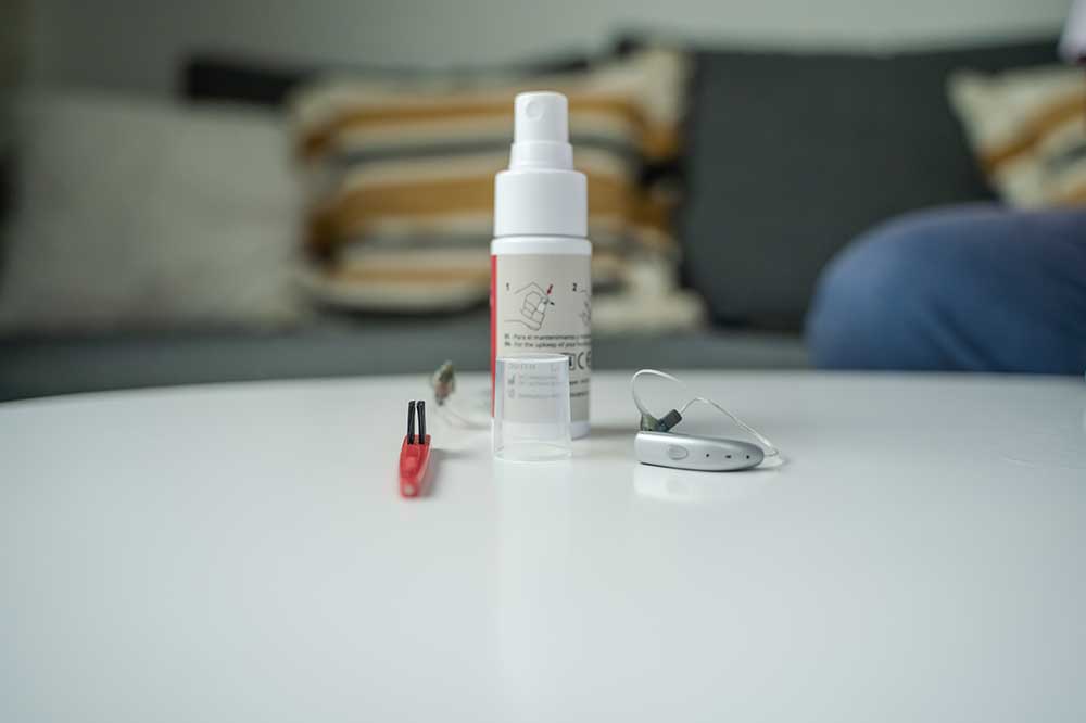 Hearing aid cleaning spray, red brush, and behind-the-ear hearing aid on a white table, for care tips from High Point Audiological in High Point, NC
