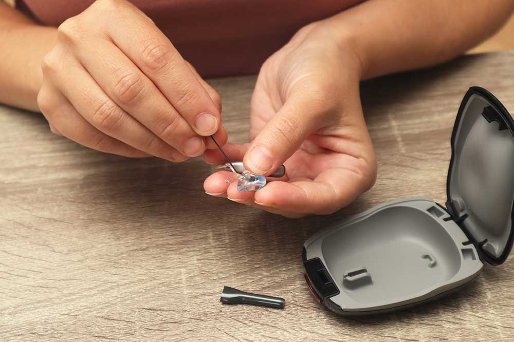 Hands using a wax loop on a hearing aid dome beside an open case, showing hearing aid cleaning tips from HEAR Ontario clinics in Ontario