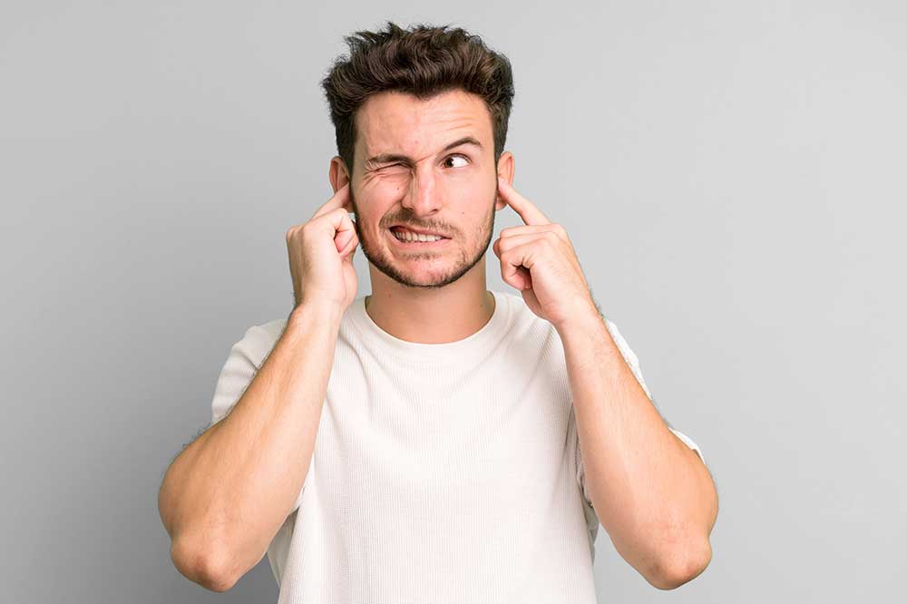 Young man grimacing and plugging his ears with his fingers, as when one hearing aid sounds too sharp, from North Hills Hearing in Hurst, TX