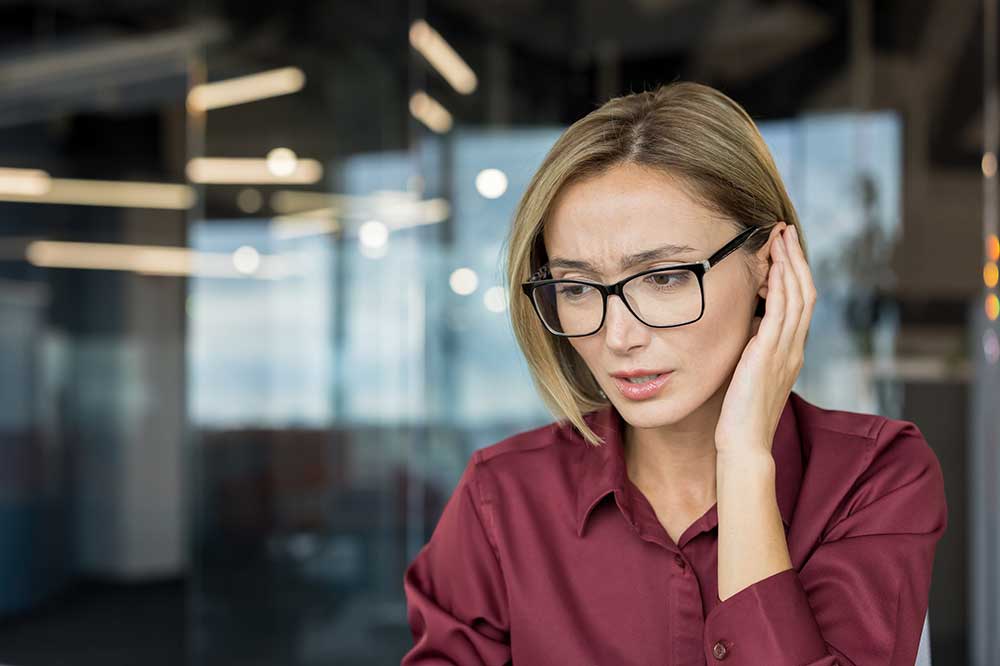Woman in glasses touching her ear with a worried look, as when one hearing aid sounds different, from High Point Audiological in High Point, NC