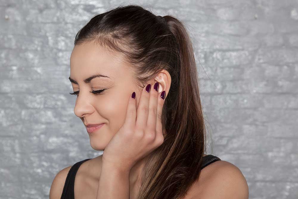 Woman with a ponytail touching her ear over a clogged, full feeling, from Colorado Ear Care, serving the Denver area and Greeley, CO
