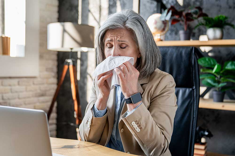 Gray-haired woman blowing her nose at her laptop desk, dealing with another sinus infection, from Southern California Ear, Nose & Throat in Long Beach